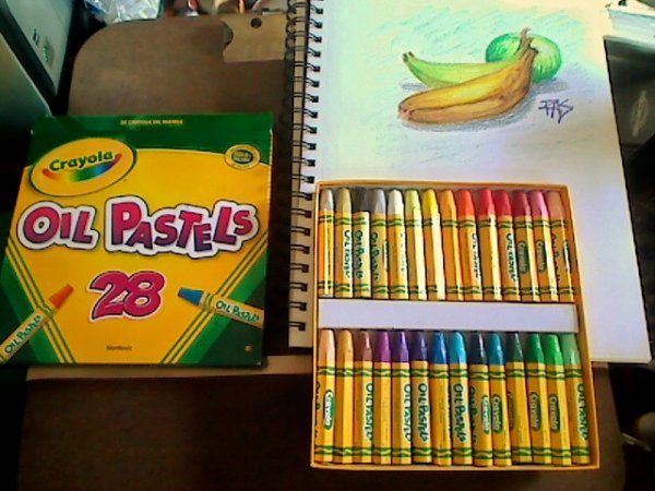 Photo of Crayola oil pastels with box lid and sketchbook with drawing of bananas and green apple.